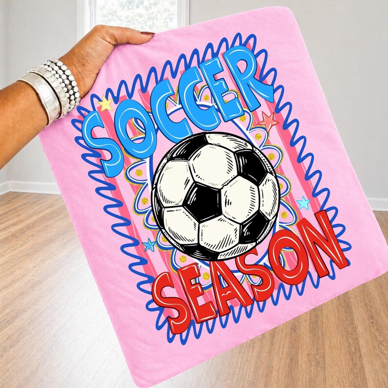 Puede incluir: Manta de f&uacute;tbol rosa con un bal&oacute;n de f&uacute;tbol en blanco y negro. La manta tiene las palabras "SOCCER SEASON" en azul y rojo. La manta est&aacute; sostenida por una persona.