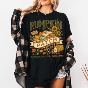 Puede incluir: Una camiseta negra con un gr&aacute;fico de un carro de madera lleno de calabazas, girasoles y un letrero que dice FRESH PICKED. Una pancarta sobre el carro dice PUMPKIN PATCH, con texto debajo que indica HAYRIDES, APPLE CIDER y HARVEST DAYS.