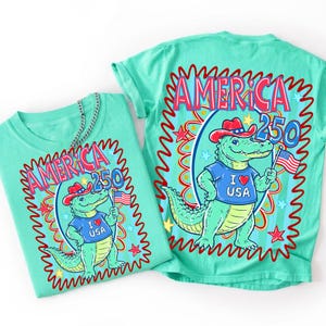 Puede incluir: Dos camisetas turquesas con un gr&aacute;fico de un caim&aacute;n con sombrero de vaquero y una camiseta con "I ❤️ USA". Las camisetas muestran el texto "AMERICA 250" en un dise&ntilde;o rojo y azul.