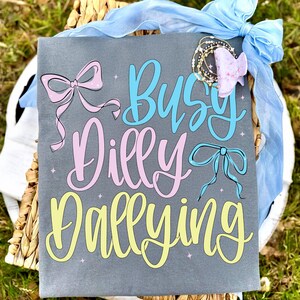 Puede incluir: Una camiseta gris con las palabras "Busy Dilly Dallying" en escritura azul, rosa y amarilla. La camiseta presenta ilustraciones de lazos en rosa y azul. Tambi&eacute;n se ven una cinta azul y una pinza para el pelo.
