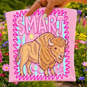 May include: Pink t-shirt with the word "SMART" in bubble letters above a cartoon bison. The bison is tan with a shaggy mane and horns. The shirt has a wavy pink border and blue and pink stripes.