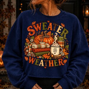 Puede incluir: Una sudadera azul marino de cuello redondo con un estampado gr&aacute;fico colorido que incluye el texto "SWEATER WEATHER". El dise&ntilde;o muestra una calabaza naranja grande, una pila de jers&eacute;is de punto doblados, un termo y una taza con el texto "GOOD THINGS AHEAD".