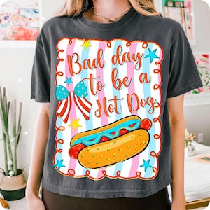 May include: Dark gray t-shirt featuring a colorful graphic with the text "Bad day to be a Hot Dog." The design includes a hot dog, bow, stars, and a striped background in red, white, and blue.