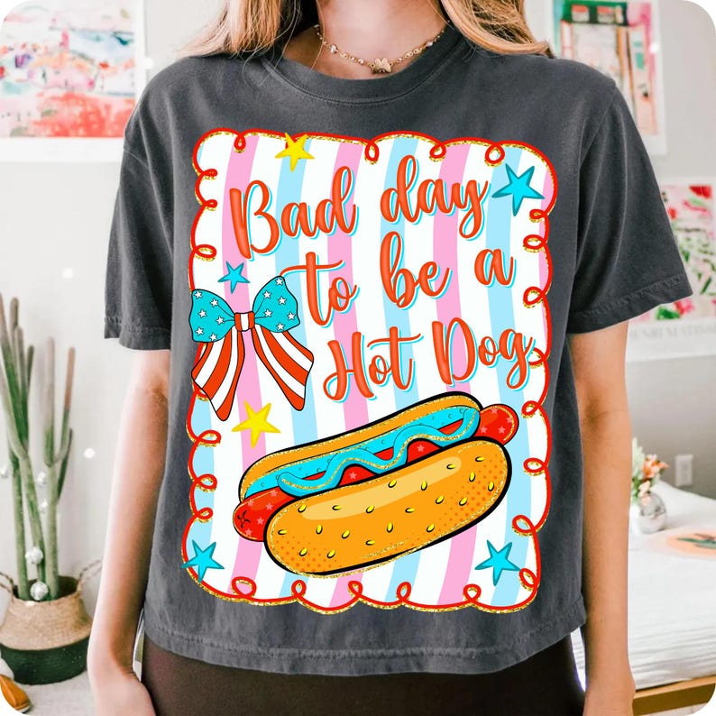 May include: Dark gray t-shirt featuring a colorful graphic with the text "Bad day to be a Hot Dog." The design includes a hot dog, bow, stars, and a striped background in red, white, and blue.
