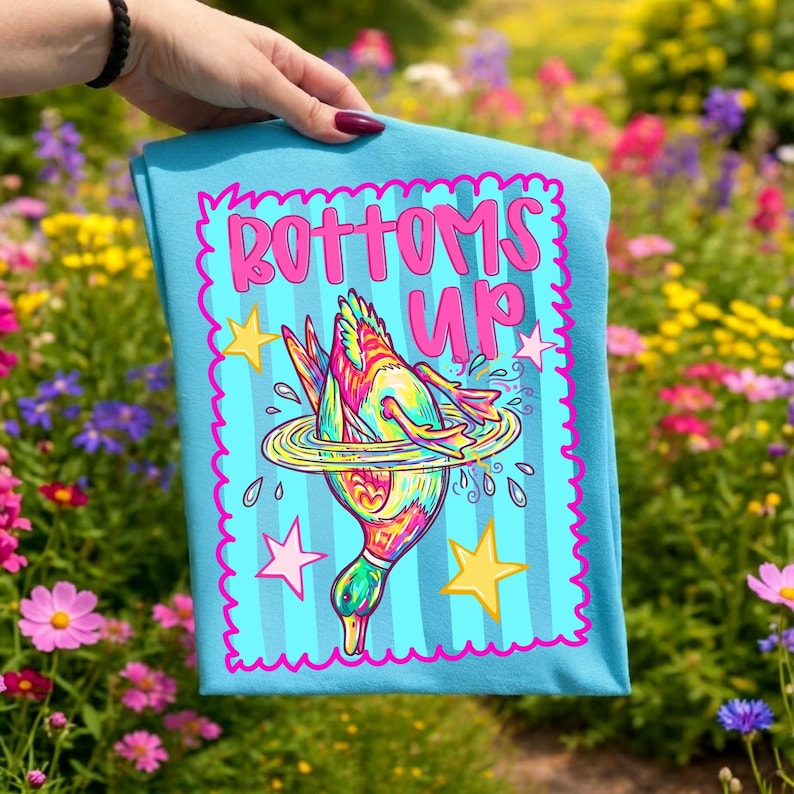 May include: A turquoise t-shirt featuring the phrase "BOTTOMS UP" in pink, with a cartoon duck diving into water. The duck is brightly coloured with yellow, pink, and green details. The design is framed by a pink wavy border and stars.