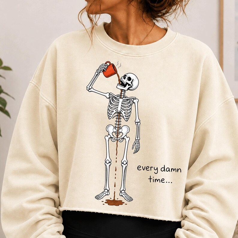 May include: A cream-coloured crewneck sweatshirt featuring a graphic of a skeleton drinking from an orange mug. The coffee flows through the skeleton's ribcage and out of the pelvic area onto the floor. The text reads, every damn time...