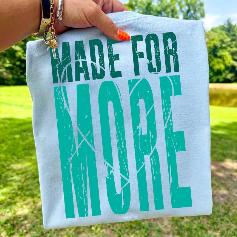 May include: A light blue t-shirt with the words "MADE FOR MORE" printed in a distressed teal font. The shirt is held up against a background of green grass and trees.