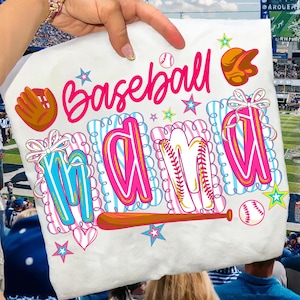Peut inclure: T-shirt blanc avec l'inscription "Baseball Mama" en rose. Le mot "Mama" est écrit en lettres majuscules avec un dessin de batte et de balle de baseball. Le t-shirt présente également des graphiques sur le thème du baseball.