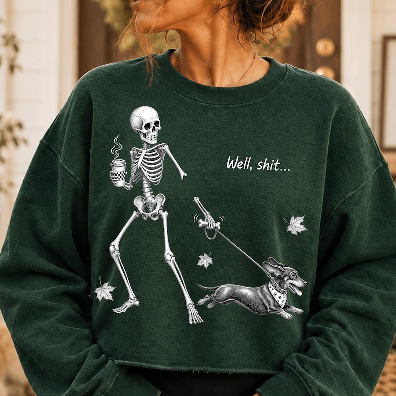 May include: A dark green crewneck sweatshirt featuring a graphic of a skeleton holding a coffee cup and walking a dachshund on a leash. The text "Well, shit..." is printed above the dog, accompanied by small white leaf illustrations.