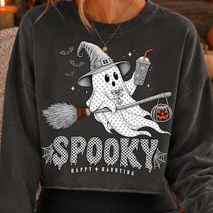 May include: A charcoal grey long-sleeve sweatshirt featuring a graphic of a ghost wearing a witch hat, riding a broomstick, and holding a drink. The ghost is decorated with stars and a pumpkin. The text reads SPOOKY HAPPY HAUNTING in a checkered font.