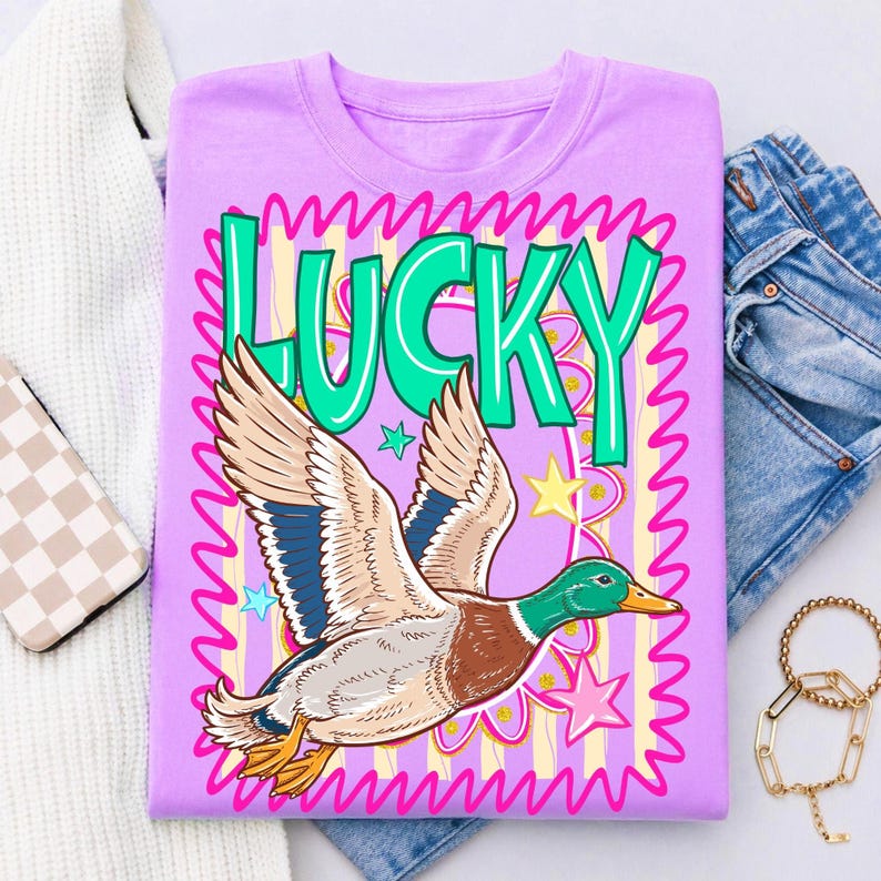 May include: A lilac t-shirt featuring a cartoon duck in flight, with the word "LUCKY" in green. The design includes stars and a pink wavy border. The shirt is displayed with jeans and a white sweater.