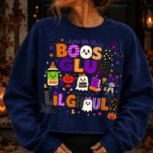 K&ouml;nnte beinhalten: Ein lang&auml;rmeliges, marineblaues Sweatshirt mit einer bunten Halloween-Grafik und dem Text "here for the BOOS & GLUE with my LILGHOULS". Das Design zeigt verschiedene Comic-Figuren wie eine Klebeflasche, ein Gespenst und eine Katze.