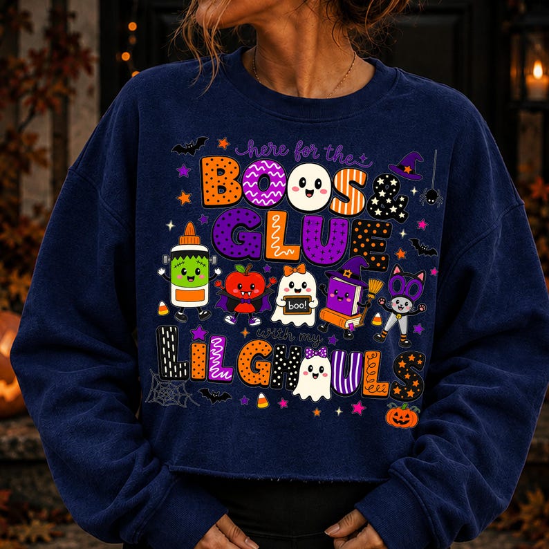 K&ouml;nnte beinhalten: Ein lang&auml;rmeliges, marineblaues Sweatshirt mit einer bunten Halloween-Grafik und dem Text "here for the BOOS & GLUE with my LILGHOULS". Das Design zeigt verschiedene Comic-Figuren wie eine Klebeflasche, ein Gespenst und eine Katze.
