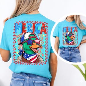 May include: A light blue t-shirt featuring a cartoon duck wearing sunglasses, a hat, and a bandana with a patriotic theme. The word "MERICA" is printed above the duck in blue and red letters.