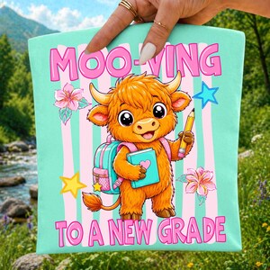 May include: A light mint green tote bag featuring a cartoon highland cow with orange fur, wearing a pink backpack, holding a blue book and a yellow pencil. The text reads MOO-VING TO A NEW GRADE, surrounded by pink flowers, a yellow star, and a blue star.