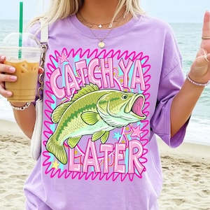 Op de afbeelding: Een lichtpaars T-shirt met een afbeelding van een groen en gele vis met open mond en de woorden "CATCH YA LATER" in roze. Het ontwerp is omlijnd met een golvend roze en blauw patroon met sterren.