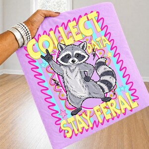 May include: A light purple blanket with a cartoon raccoon illustration. The raccoon is gray and white, giving a rock and roll hand gesture. Text reads "COLLECT DATA STAY FERAL" in yellow and pink.