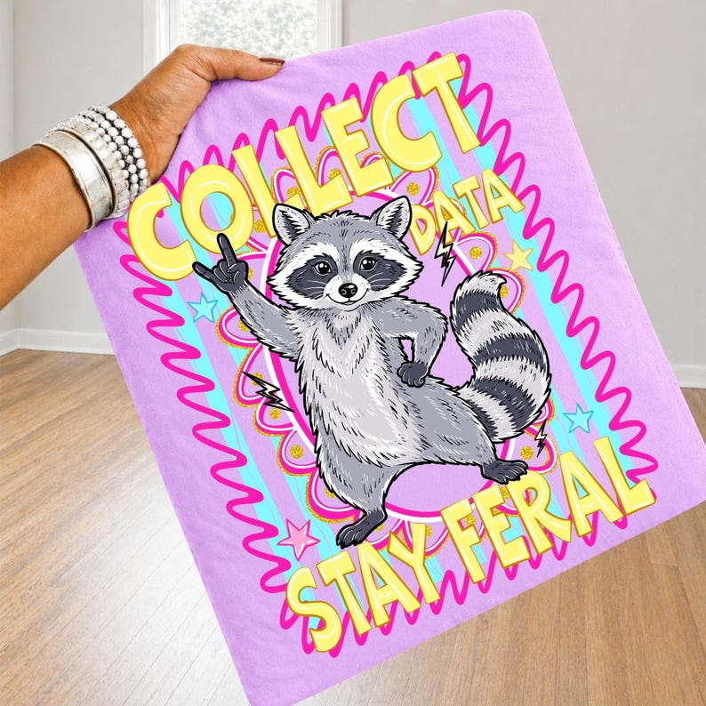May include: A light purple blanket with a cartoon raccoon illustration. The raccoon is gray and white, giving a rock and roll hand gesture. Text reads "COLLECT DATA STAY FERAL" in yellow and pink.