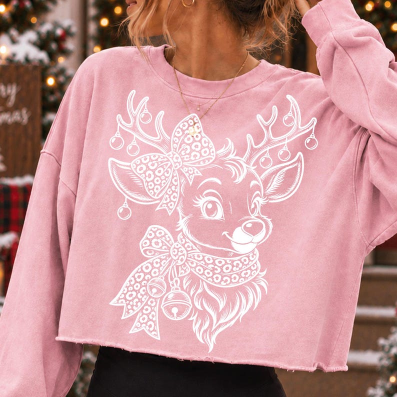 May include: A light pink cropped sweatshirt featuring a white graphic print of a reindeer with a leopard print bow and scarf. The reindeer has antlers decorated with small hanging ornaments. The sweatshirt has long sleeves and a raw hemline.