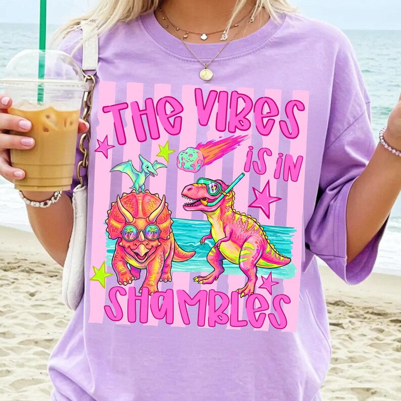 Peut inclure: T-shirt lilas avec un motif graphique color&eacute;. Le design comprend des dinosaures de dessins anim&eacute;s portant des lunettes de soleil et un tuba, avec le texte "The Vibes is in Shambles" en rose. Le t-shirt a une coupe ample.