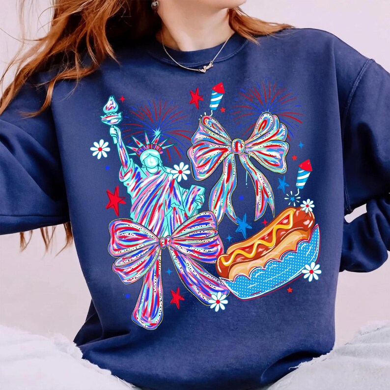May include: Navy blue sweatshirt featuring a patriotic design. The graphic includes the Statue of Liberty, bows, fireworks, a hot dog, and stars. The design is in red, white, and blue colors, perfect for celebrating Independence Day.