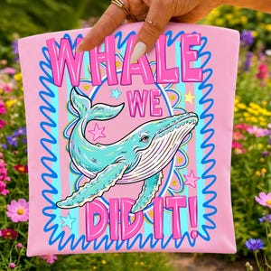 K&ouml;nnte beinhalten: Rosafarbenes T-Shirt mit einer t&uuml;rkisfarbenen Walillustration und dem Text "WHALE WE DID IT!" in Pink. Das Design umfasst Sterne und einen blau-t&uuml;rkisfarbenen gestreiften Hintergrund. Das T-Shirt wird von einer Hand gehalten.