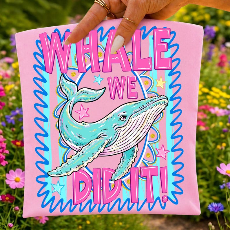 K&ouml;nnte beinhalten: Rosafarbenes T-Shirt mit einer t&uuml;rkisfarbenen Walillustration und dem Text "WHALE WE DID IT!" in Pink. Das Design umfasst Sterne und einen blau-t&uuml;rkisfarbenen gestreiften Hintergrund. Das T-Shirt wird von einer Hand gehalten.