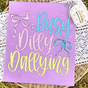 Puede incluir: Una camiseta de color lila con la frase "Busy Dilly Dallying" en escritura pastel azul, rosa y amarillo, con ilustraciones de lazos. Un peque&ntilde;o bolso floral con una cadena es visible en la esquina superior derecha.