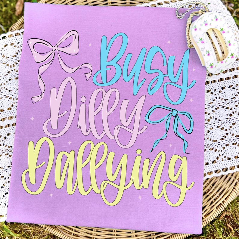 Puede incluir: Una camiseta de color lila con la frase "Busy Dilly Dallying" en escritura pastel azul, rosa y amarillo, con ilustraciones de lazos. Un peque&ntilde;o bolso floral con una cadena es visible en la esquina superior derecha.