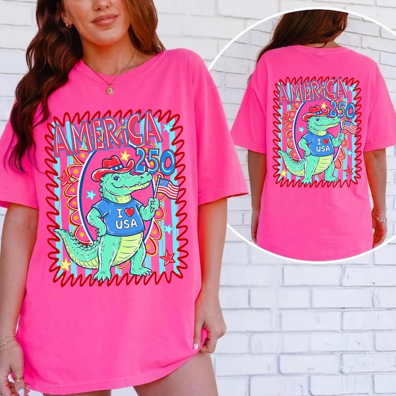 Puede incluir: Una camiseta rosa brillante con un cocodrilo de dibujos animados que lleva un sombrero de vaquero y una camiseta "I Love USA", sosteniendo una peque&ntilde;a bandera americana. La camiseta tiene el texto "AMERICA 250" en un marco decorativo.