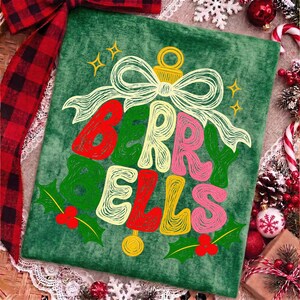 May include: A green velvet fabric square featuring a graphic of a Christmas ornament shaped like the text BERRY BELLS. The design includes a white line-art bow, yellow stars, and red holly berries with green leaves on a textured green background.