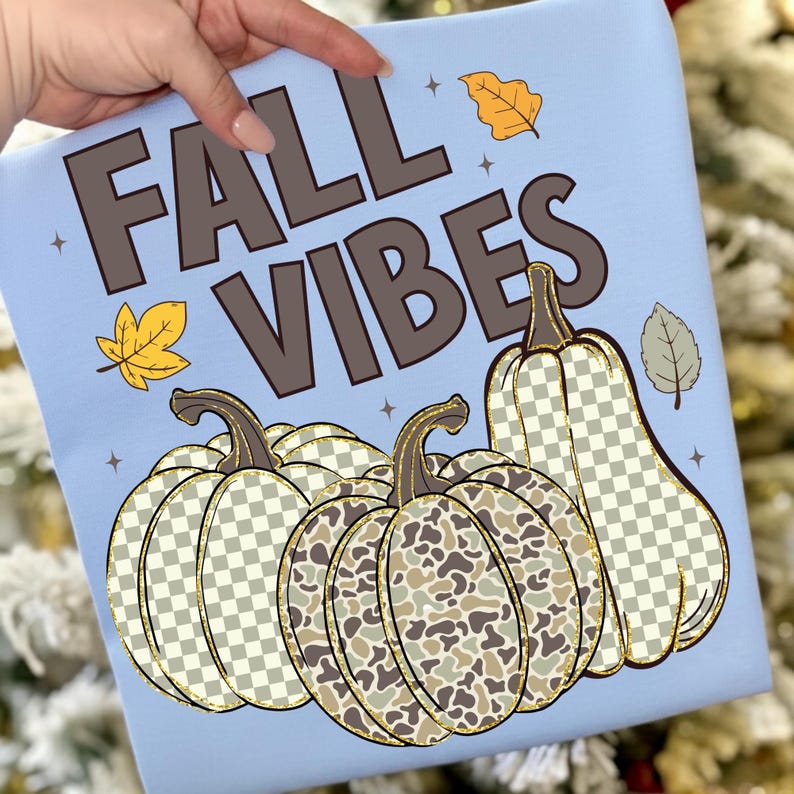 Puede incluir: Una sudadera azul claro con el texto marr&oacute;n FALL VIBES, acompa&ntilde;ada de tres calabazas con patrones de cuadros y camuflaje, acentuada por peque&ntilde;as ilustraciones de hojas amarillas y grises y diminutas estrellas de cuatro puntas.
