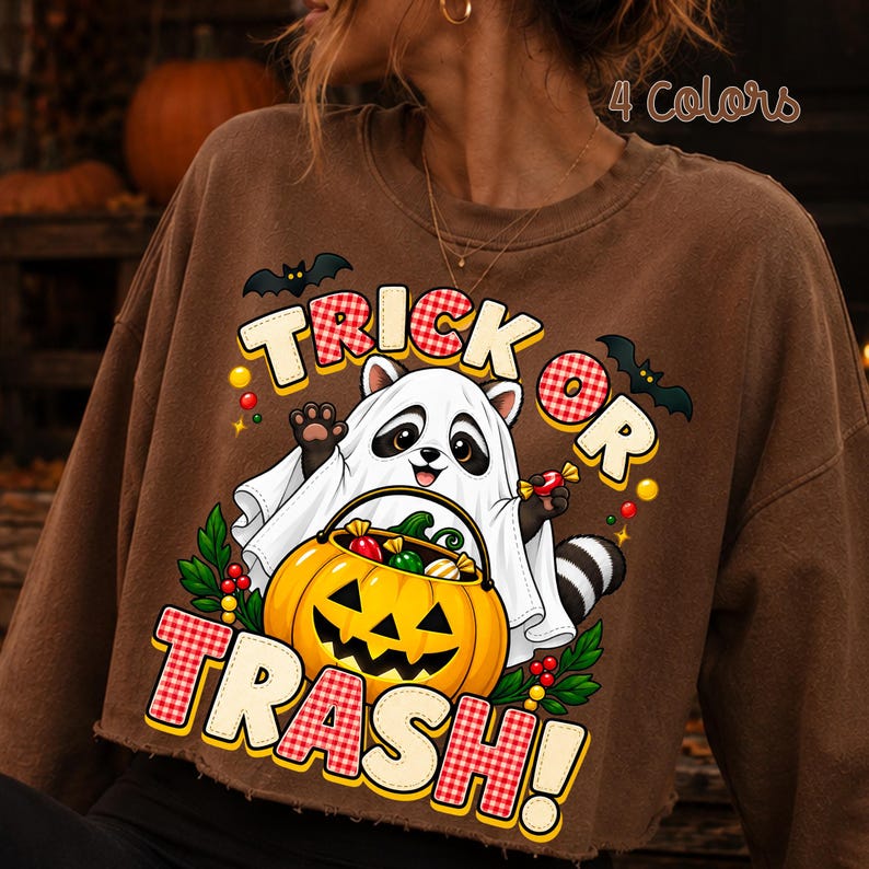 May include: A brown crewneck sweatshirt featuring a graphic of a raccoon dressed as a ghost holding a jack-o'-lantern candy bucket. The text reads "TRICK OR TRASH!" in red and white checkered lettering, surrounded by bats, holly, and colourful candy.