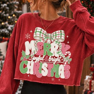 K&ouml;nnte beinhalten: Ein rotes, lang&auml;rmeliges bauchfreies Sweatshirt mit einer gro&szlig;en Grafik einer gr&uuml;n-wei&szlig; karierten Schleife &uuml;ber dem Schriftzug Merry joyful & bright Christmas in wei&szlig;er, gr&uuml;ner und roter Schrift, verziert mit Stechpalmen und einer Zuckerstange.