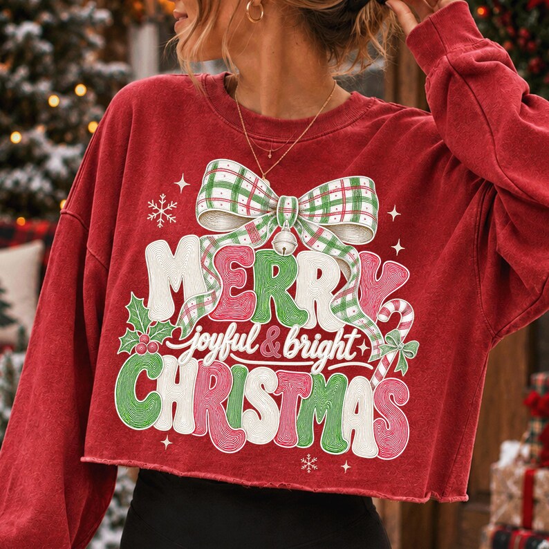 K&ouml;nnte beinhalten: Ein rotes, lang&auml;rmeliges bauchfreies Sweatshirt mit einer gro&szlig;en Grafik einer gr&uuml;n-wei&szlig; karierten Schleife &uuml;ber dem Schriftzug Merry joyful & bright Christmas in wei&szlig;er, gr&uuml;ner und roter Schrift, verziert mit Stechpalmen und einer Zuckerstange.