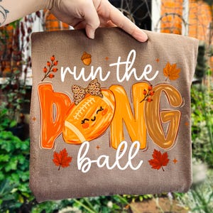 Puede incluir: Una camiseta marr&oacute;n con el texto "run the ball" en letra cursiva blanca. El gr&aacute;fico incluye un bal&oacute;n de f&uacute;tbol americano naranja con un lazo de estampado de leopardo y una cara sonriente, rodeado de hojas de oto&ntilde;o, bayas y una bellota.