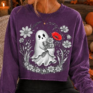 K&ouml;nnte beinhalten: Ein violettes Langarm-Sweatshirt mit Rundhalsausschnitt und einer Grafik eines wei&szlig;en Geistes, der einen Blumenstrau&szlig; h&auml;lt. Das Design zeigt schwarz-wei&szlig;e Strichzeichnungen von Blumen um den Geist herum, mit einer einzelnen roten Mohnblume.