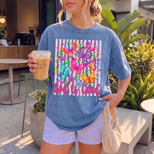 May include: A blue short-sleeve t-shirt featuring a graphic print of colorful butterflies over a white and blue striped background. The text "God is Good" is printed in pink cursive script at the bottom of the rectangular graphic design.