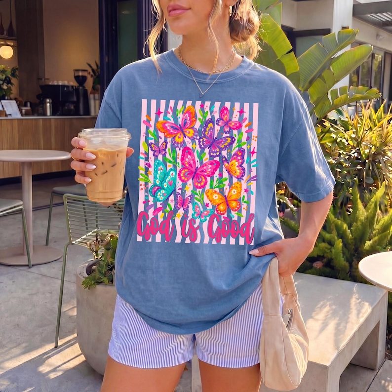 May include: A blue short-sleeve t-shirt featuring a graphic print of colorful butterflies over a white and blue striped background. The text "God is Good" is printed in pink cursive script at the bottom of the rectangular graphic design.