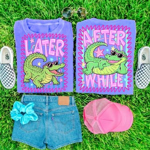 May include: A flat lay featuring two purple t-shirts with alligator graphics and the phrases "LATER" and "AFTER WHILE". The image also includes denim shorts, a pink trucker hat, a blue scrunchie, sunglasses, and checkered slip-on shoes.
