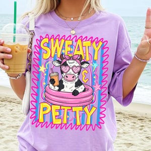 May include: A lilac-colored t-shirt with the words "SWEATY PETTY" in yellow and pink. The graphic features a cow wearing heart-shaped sunglasses and a pink headband, holding a drink while floating in a pink inner tube.