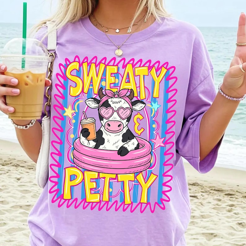 May include: A lilac-colored t-shirt with the words "SWEATY PETTY" in yellow and pink. The graphic features a cow wearing heart-shaped sunglasses and a pink headband, holding a drink while floating in a pink inner tube.