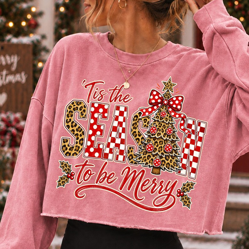 Op de afbeelding: Een oudroze cropped sweatshirt met lange mouwen en een grafische print met de tekst 'Tis the SEASON to be Merry'. Het ontwerp bevat een kerstboom met luipaardprint, een rood-witte strik met stippen, hulstbladeren en rode bessen.