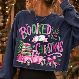 May include: A navy blue long-sleeve sweatshirt featuring a graphic print with the text "BOOKED for CHRISTMAS". The design includes a pink pickup truck carrying a stack of books and a Christmas tree, accented with a pink bow and green foliage.