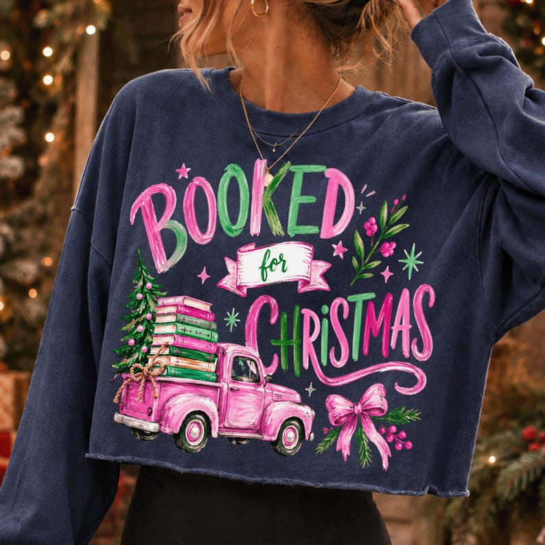 May include: A navy blue long-sleeve sweatshirt featuring a graphic print with the text "BOOKED for CHRISTMAS". The design includes a pink pickup truck carrying a stack of books and a Christmas tree, accented with a pink bow and green foliage.
