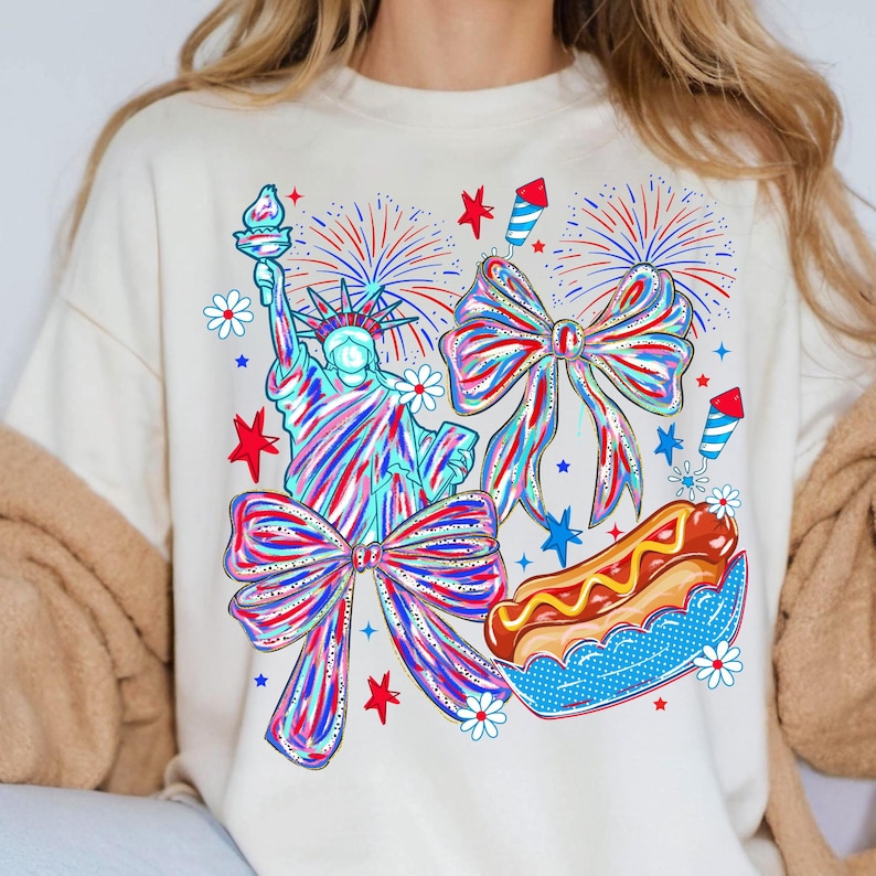 May include: Cream-colored sweatshirt featuring a patriotic design. The graphic includes the Statue of Liberty, bows, fireworks, a hot dog, and stars in red, white, and blue. The design is a colorful illustration.