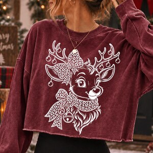 May include: A maroon long-sleeve cropped sweatshirt featuring a white graphic print of a reindeer. The reindeer has a leopard print bow on its head and a matching leopard print scarf with bells around its neck, with ornaments hanging from its antlers.