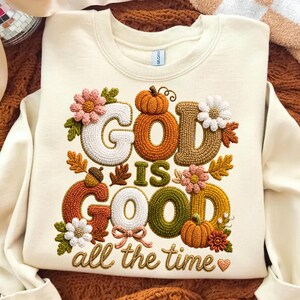 Pu&ograve; includere: Una felpa girocollo color crema con un disegno ricamato che riporta il testo "GOD IS GOOD all the time". Le lettere sono riempite con punti testurizzati multicolori e circondate da zucche, foglie autunnali e fiori.