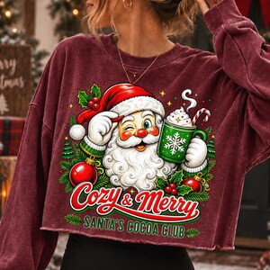 K&ouml;nnte beinhalten: Ein bordeauxrotes Langarm-Sweatshirt mit einer Grafik eines zwinkernden Weihnachtsmanns, der einen gr&uuml;nen Kakaobecher mit Zuckerstange h&auml;lt. Der Text lautet Cozy & Merry SANTA'S COCOA CLUB, umgeben von Stechpalmen, Tannen und roten Kugeln.