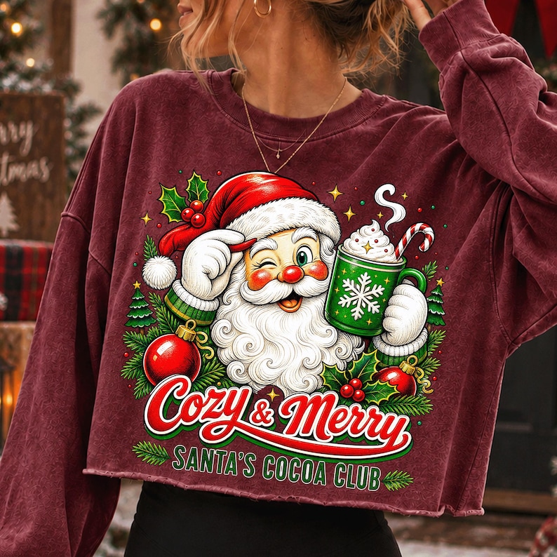 K&ouml;nnte beinhalten: Ein bordeauxrotes Langarm-Sweatshirt mit einer Grafik eines zwinkernden Weihnachtsmanns, der einen gr&uuml;nen Kakaobecher mit Zuckerstange h&auml;lt. Der Text lautet Cozy & Merry SANTA'S COCOA CLUB, umgeben von Stechpalmen, Tannen und roten Kugeln.